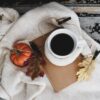 cup of coffee on top of a book, surrounded by cozy white blanket, fall leaves and a mini pumpkin